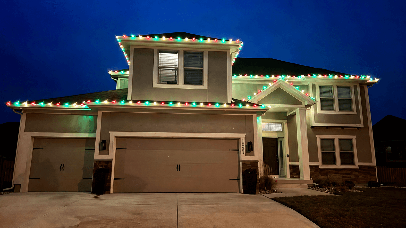 A two-story home with red, green and white holiday lights outlining the roof at dusk