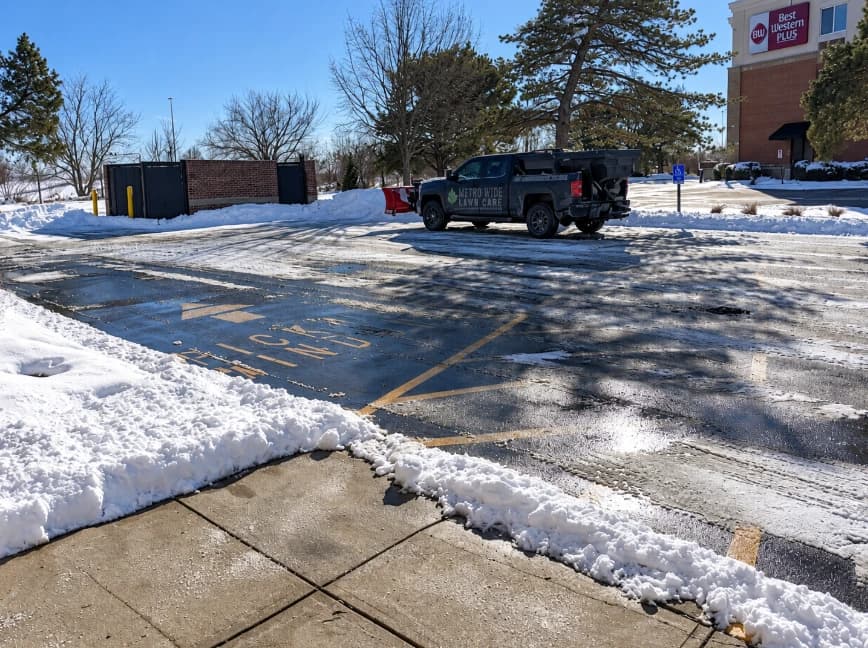Metro Wide Lawn Care truck with a snowplow and spreader in a snowy parking lot beside a cleared sidewalk